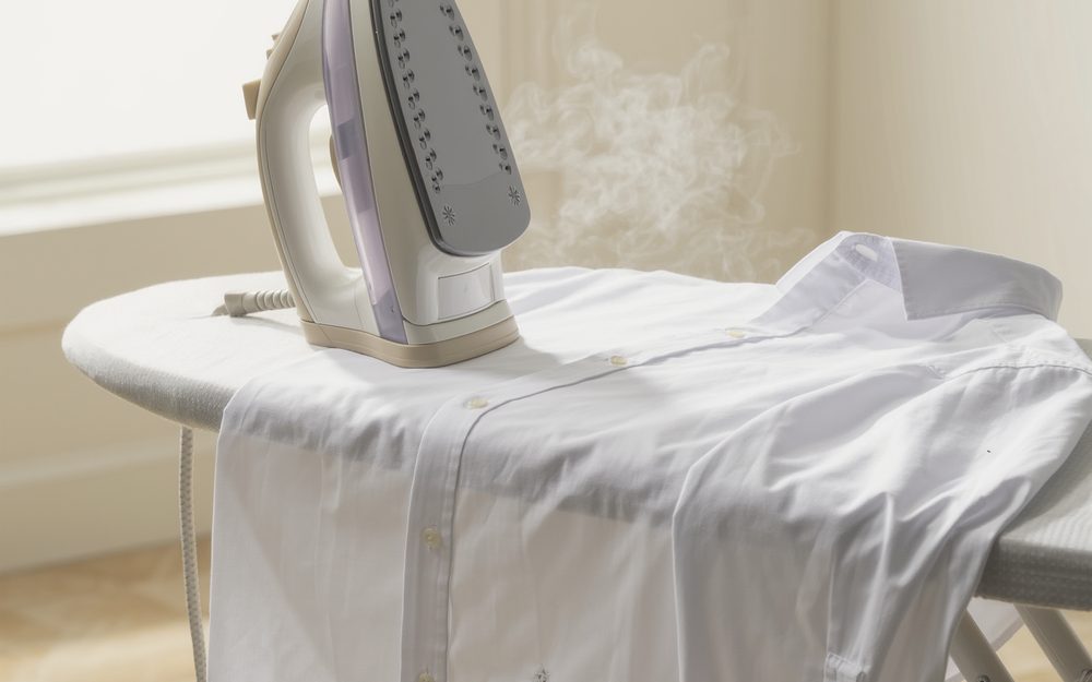 A steam iron resting on a white shirt laid over an ironing board, in front of a bright window.