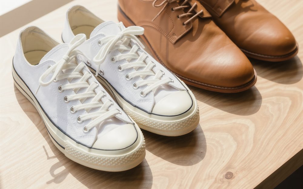A pair of white canvas sneakers and a pair of tan leather shoes on a timber sill in sunlight.