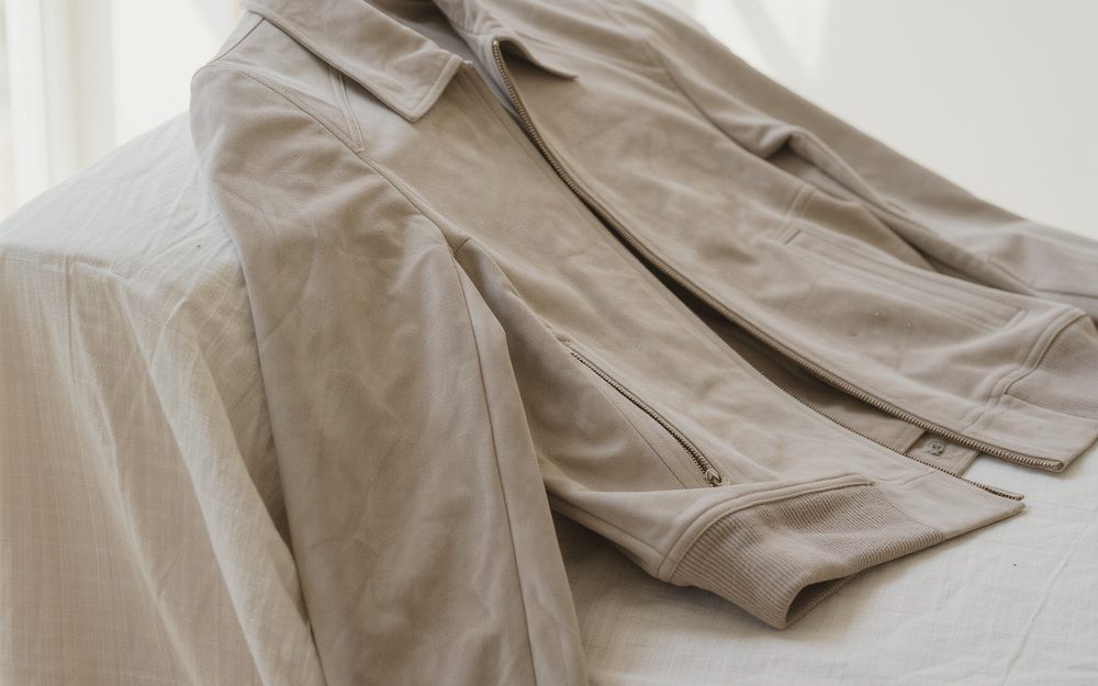 A stone-coloured jacket laid across a white bed in soft window light.